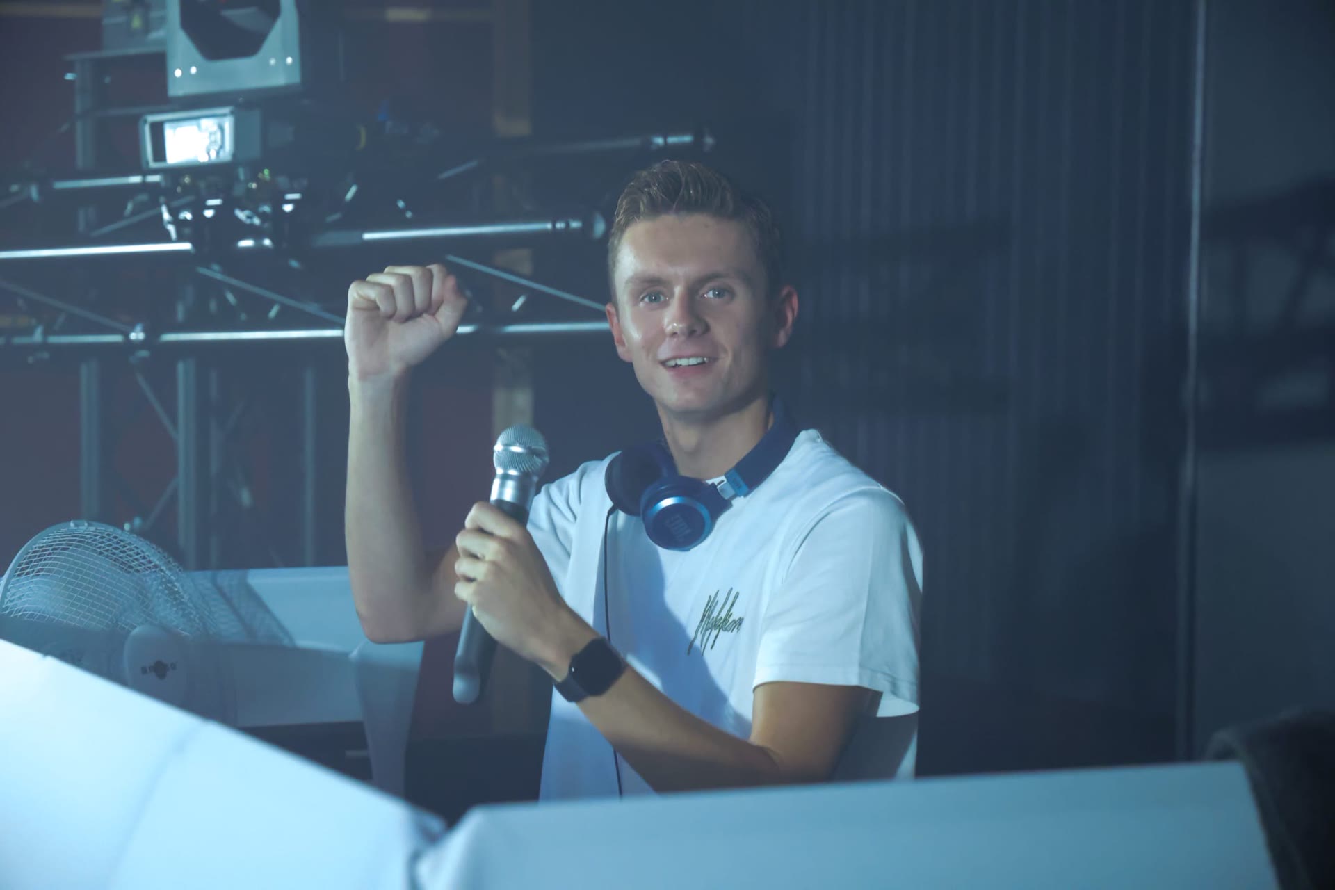 Casper met microfoon en koptelefoon achter de draaitafels, onder de truss